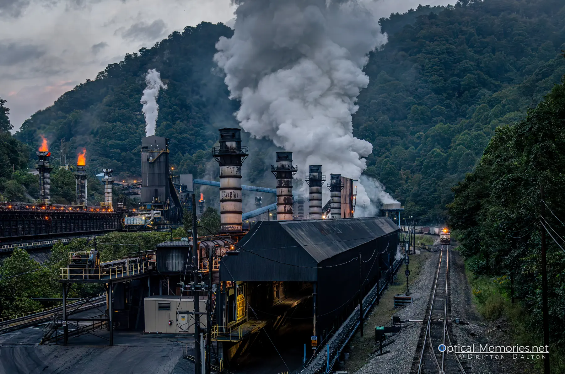 Industrial plant glowing with flame and smoke beneath a forested Appalachian ridge